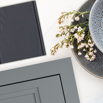 Gray painted cabinet door and finish sample laying on a white countertop. The sample shows dark navy blue painte oak with a wood texture. Explore kitchen color trends from the expert color forecasters at Dura Supreme Cabinetry.