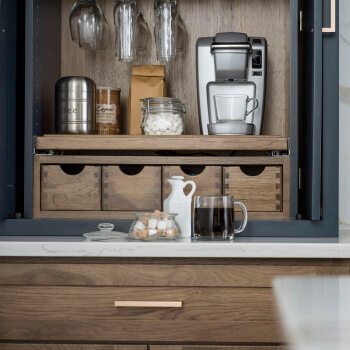 A beverage center larder cabinet from Dura Supreme Cabinetry in a Navy blue and medium stained hickory kitchen. This pantry cabinet sits on the countertop and offers organized storage and integrated features for creating an optimized coffee station that hides behind poket cabinet doors.