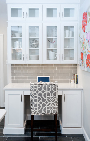 What is a kitchen layout? This is an office desk within the kitchen design with pretty white painted cabinets and glass cabinet doors.