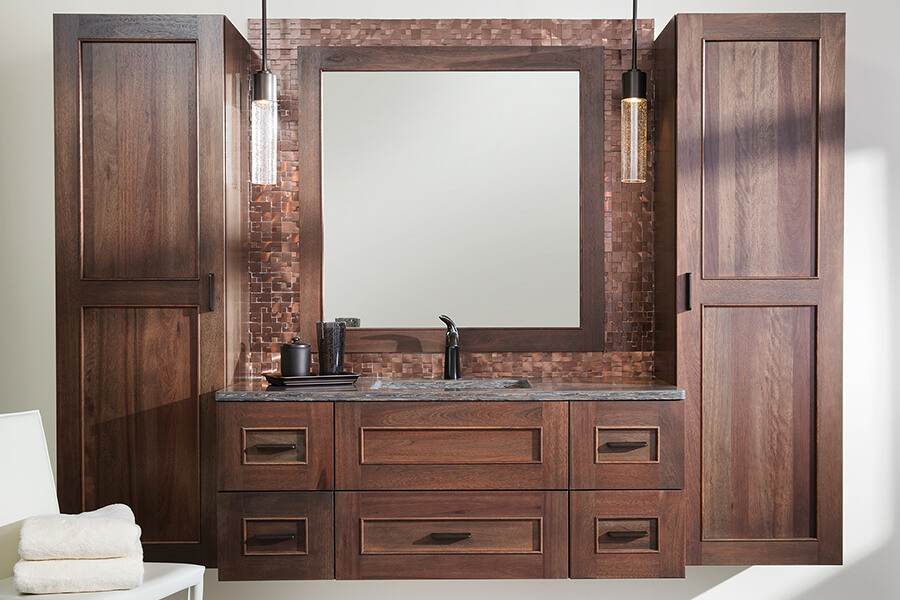 A transitional style floating bathroom vanity with wall-hung linen cabinets. A dark reddish stained wood accents the copper backsplash tiles. These bathroom cabinets are crafted with Dura Supreme's Bria Cabinetry, a frameless/full-access cabinet product line sold in the US.
