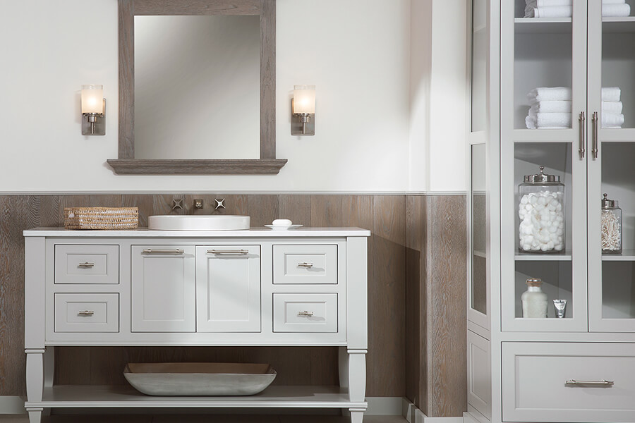 Framed Crestwood Cabinetry by Dura Supreme is used in a coastal style master bathroom on a furniture style vanity. The bathroom vanity cabinets feature inset flat panel door styles with decorative turned posts and a floor shelf for displaying decoratives.