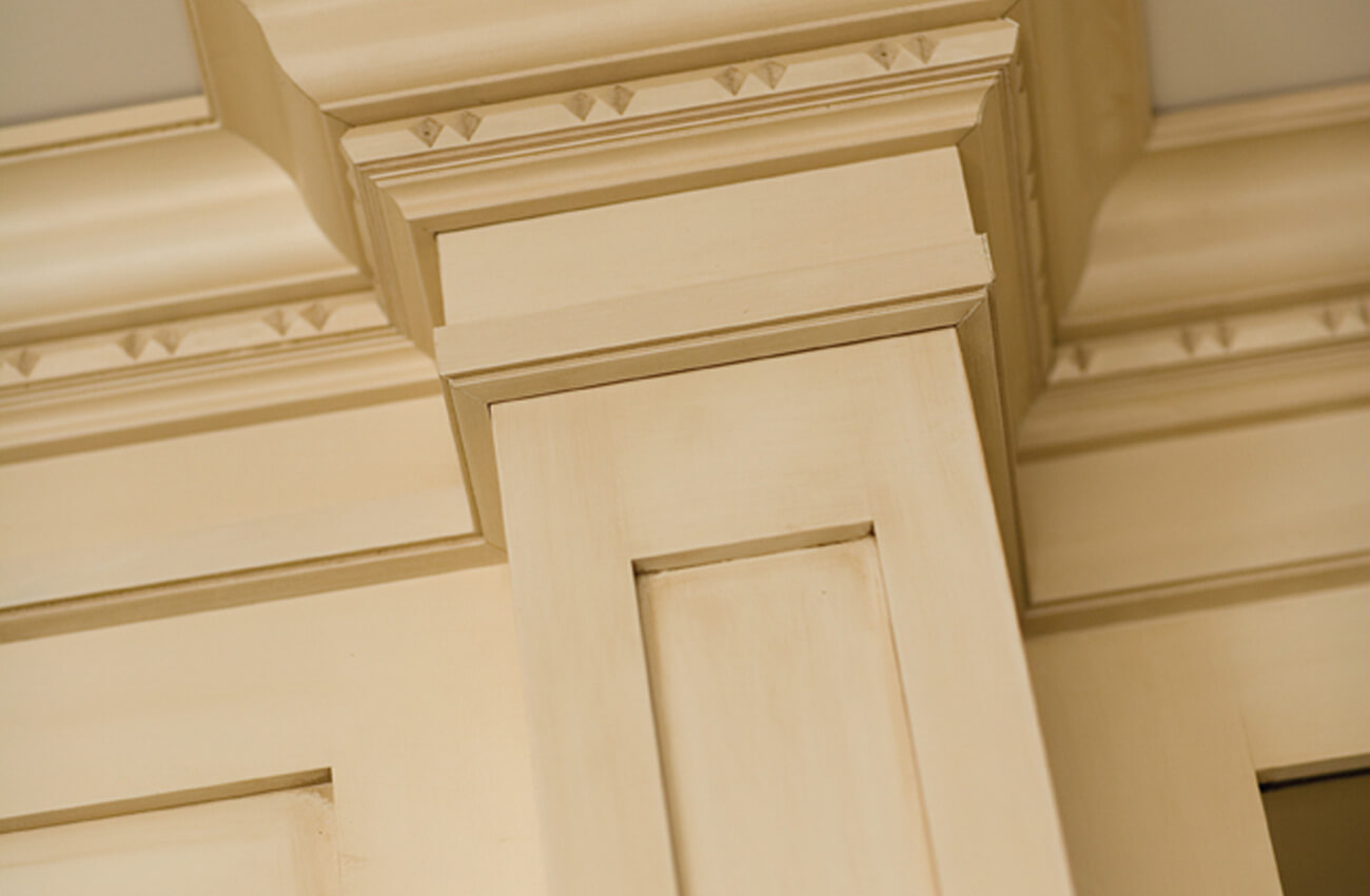 The details of a traditional style kitchen top cabinet molding. The crown molding stacks and the cabinet doors feature a warm off-white glazed paint finish. The finish artisians hand apply multiple layers of finish to create this look.