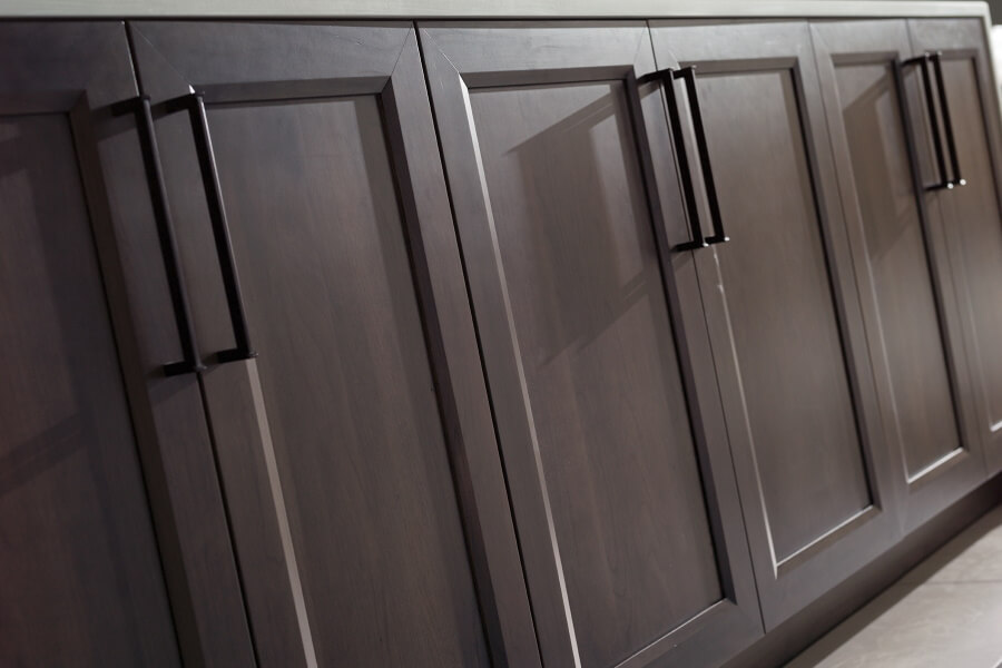 A row of cabinet doors with a dark cool-brown stain with flat panel cabinet doors. A Full Overlay door style overlaps the face frame of the cabinet by 1 1/4″, leaving a 1/4″ of the face frame exposed. Learn how to select your cabinetry construction method when shopping for kitchen cabinetry.