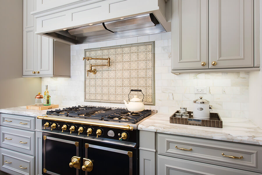 Mixed metals and finsihes add a beautiful trendy look to this kitchen design. Beautiful slide-in range with double ovens in a gray kitchen design with gray painted kitchen cabinets and light gray backsplash tiles. Gold hardware, pot filler, and accents add warmth to the cool color palette. A detailed wood hood adds elegance to the design.