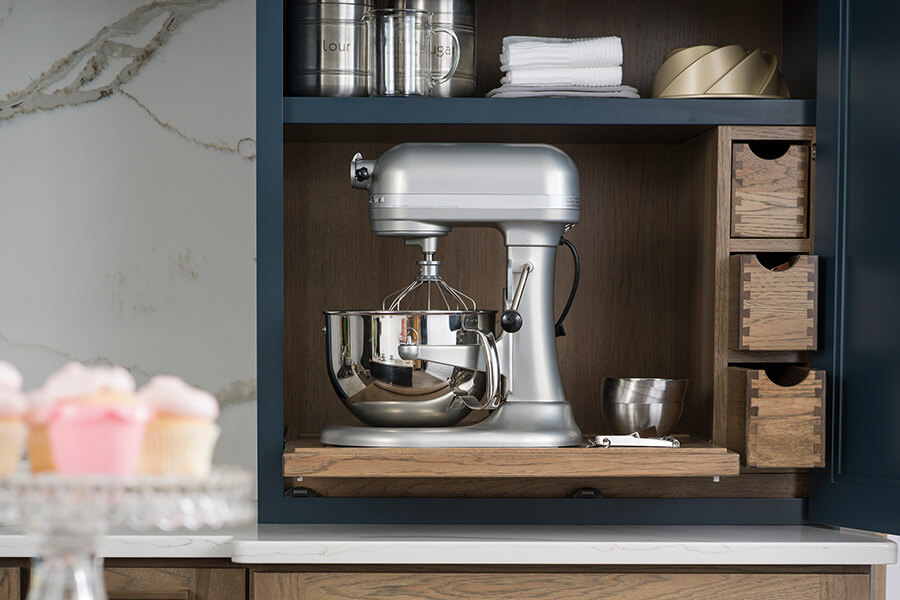 A popular kitchen design trend for storage is concealed countertop storage for small kitchen appliances and all the tools needed for using the appliance. This larder cabinet is an all-in-one baking workstation with a kitchen aid mixer and lots of storage for baking supplies. The kitchen has a navy blue and hickory color palette.