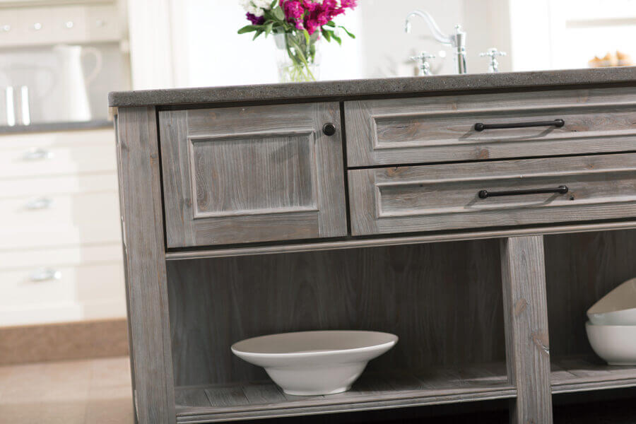 A weathered wood kitchen island cabinet with a light gray stain. This finish process looks like aged barnwood or worn driftwood. Learn about the the intricacies of kitchen cabinet construction.