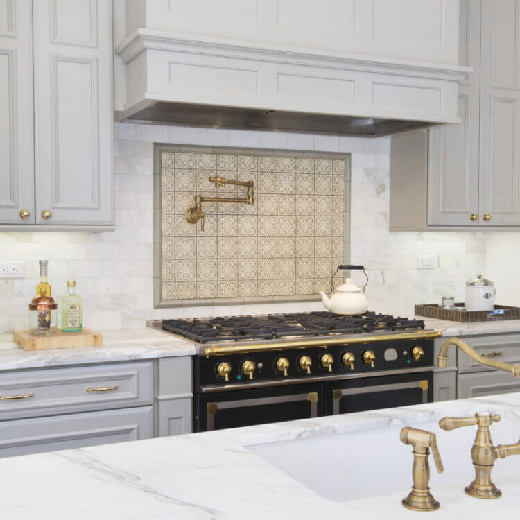 Beautiful slide-in range with double ovens in a gray kitchen design with gray painted kitchen cabinets and light gray backsplash tiles. Gold hardware, pot filler, and accents add warmth to the cool color palette. A detailed wood hood adds elegance to the design.