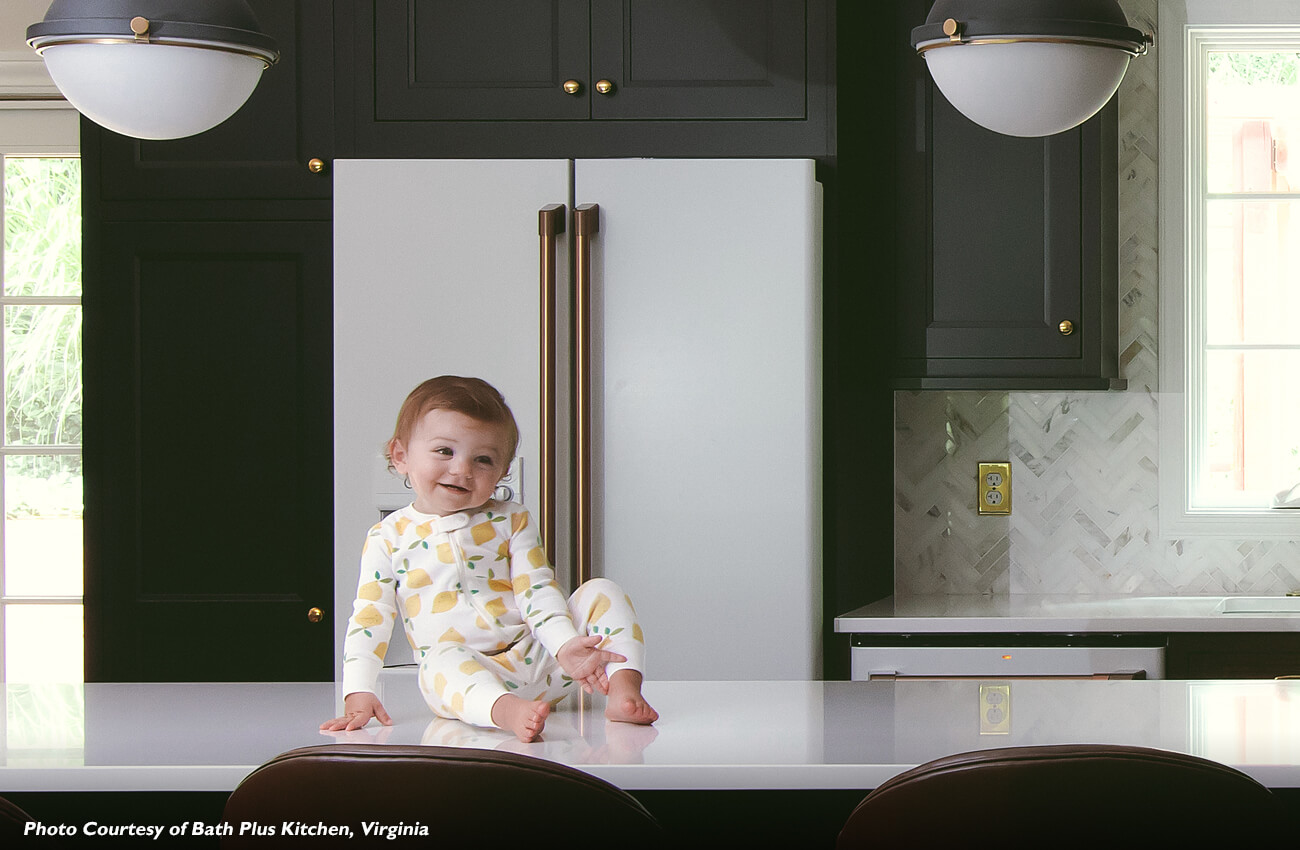 An adorable baby sitting on the kitchen island countertop in a remodeled kitchen design with dark black painted cabinets and white kitchen appliances with gold hardware and details. This trendy kitchen design is beautiful and family-friendly. The kitchen of your dreams is now a reality, and simply yours to share and enjoy. Our Kitchen Love Stories highlight the results other homeowners have enjoyed with Dura Supreme.