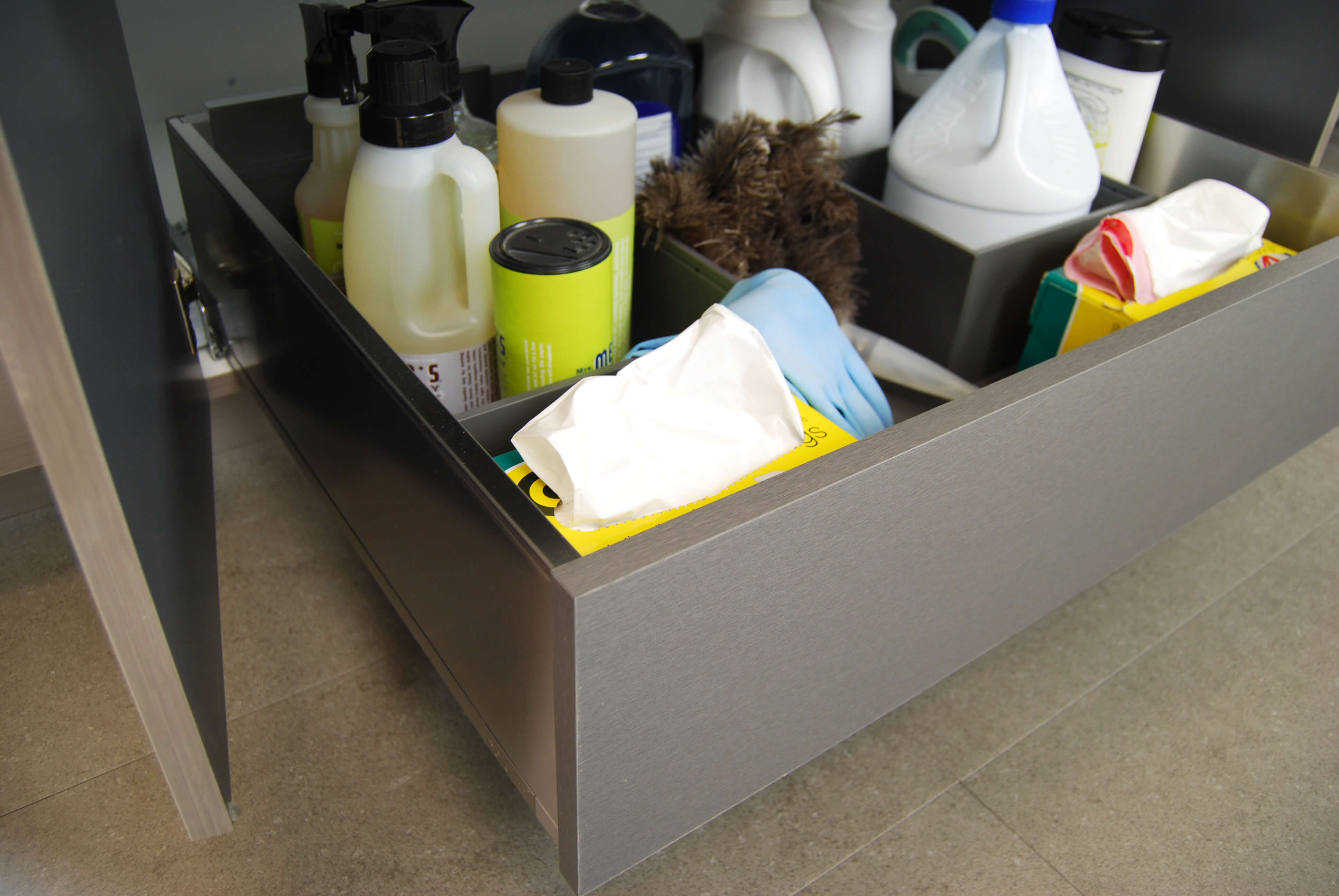 Kitchen cabinet Roll-Out shelves are shown in the Stainless Steel option and used for storing cleaning supplies under the sink in this example.