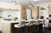 This kitchen is highlighted with a flat panel door style in an off-white painted finish mixed with a slab wood plank door with Lodge Oak texture. The kitchen island offers seating for four and has a kitchen sink that is positioned across from the cooktop.