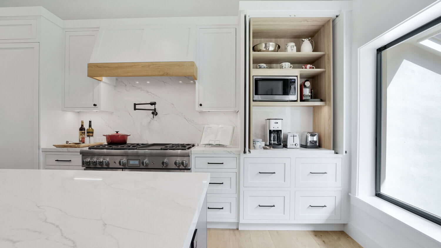 This beverage station hides all of the small appliances for crafting beverages as well as a toaster with an appliance door above the beverage fridge as well as cup storage in the cabinet to the side and pantry storage for coffee supplies and breakfast fixings in the cabinet above. The kitchen has white painted inset cabinets and a modern wood hood with a light stained wood frieze accent.