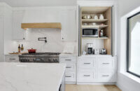 This beverage station hides all of the small appliances for crafting beverages as well as a toaster with an appliance door above the beverage fridge as well as cup storage in the cabinet to the side and pantry storage for coffee supplies and breakfast fixings in the cabinet above. The kitchen has white painted inset cabinets and a modern wood hood with a light stained wood frieze accent.