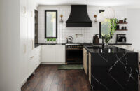 This alluring kitchen design features a skinny shaker door style with thin rails and stiles using inset cabinets. The cabinetry colors combine matte back metal glass cabinet doors, with an off-white paint, and a light stained white oak. A black painted curved hood creates a beautiful feature above the cooktop.