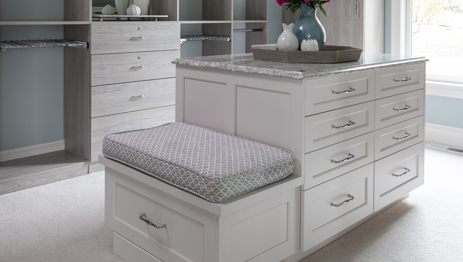 A island inside a walk-in closet that features a boot bench with seating on the end cap.