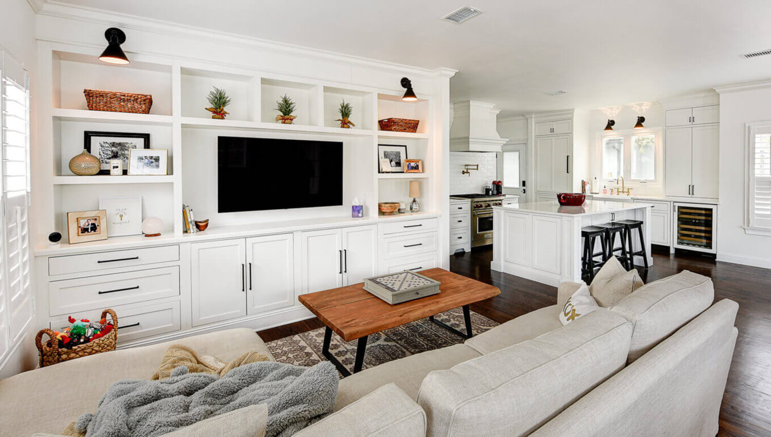 An all white open concept living room and kitchen. The built-in entertainment center matches the bright white shaker kitchen cabinetry.