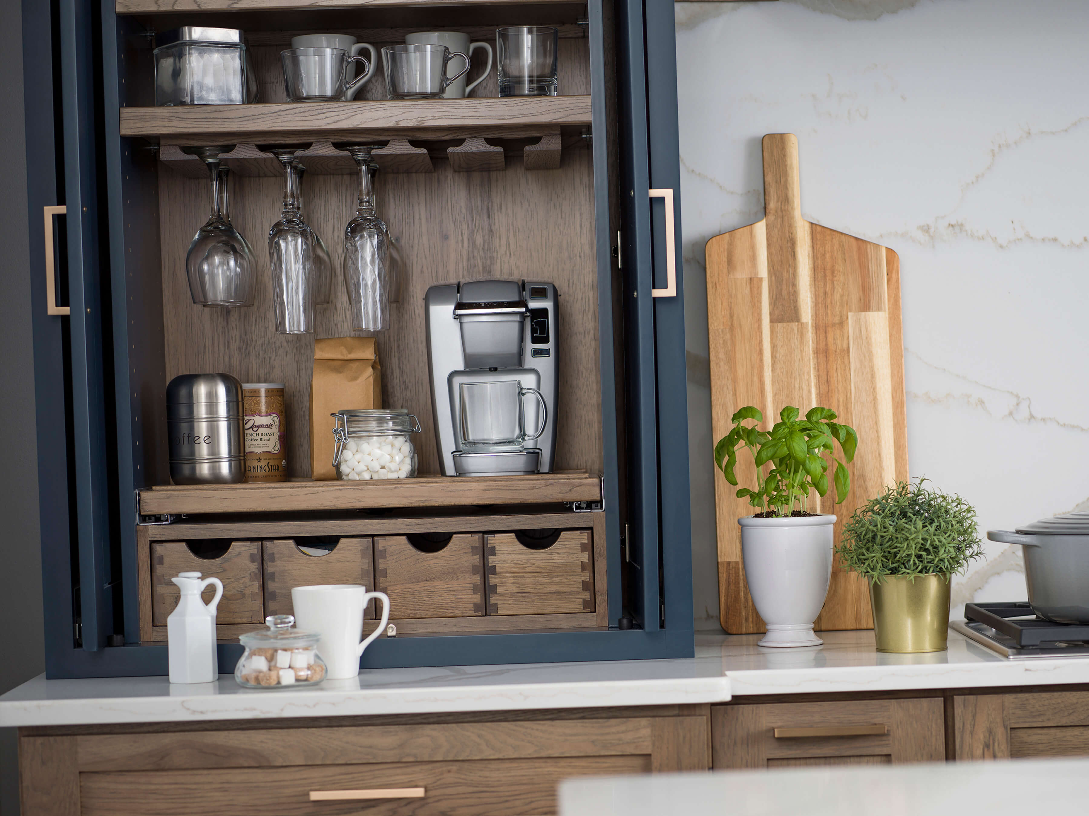 This breakfast work zone is design has everything the homeowner needs for crafting beverages for all occasions as well as a place for preparing breakfast. The larder cabinet houses a powered coffee machine, with coffee cups and wine glasses above and all the supplies for making a wide variety of beverages in the cubbies below. The apothecary drawers below hold stir sticks, tea bags, hot cocoa packets, sugar, sweeteners, and liquid creamer cups.