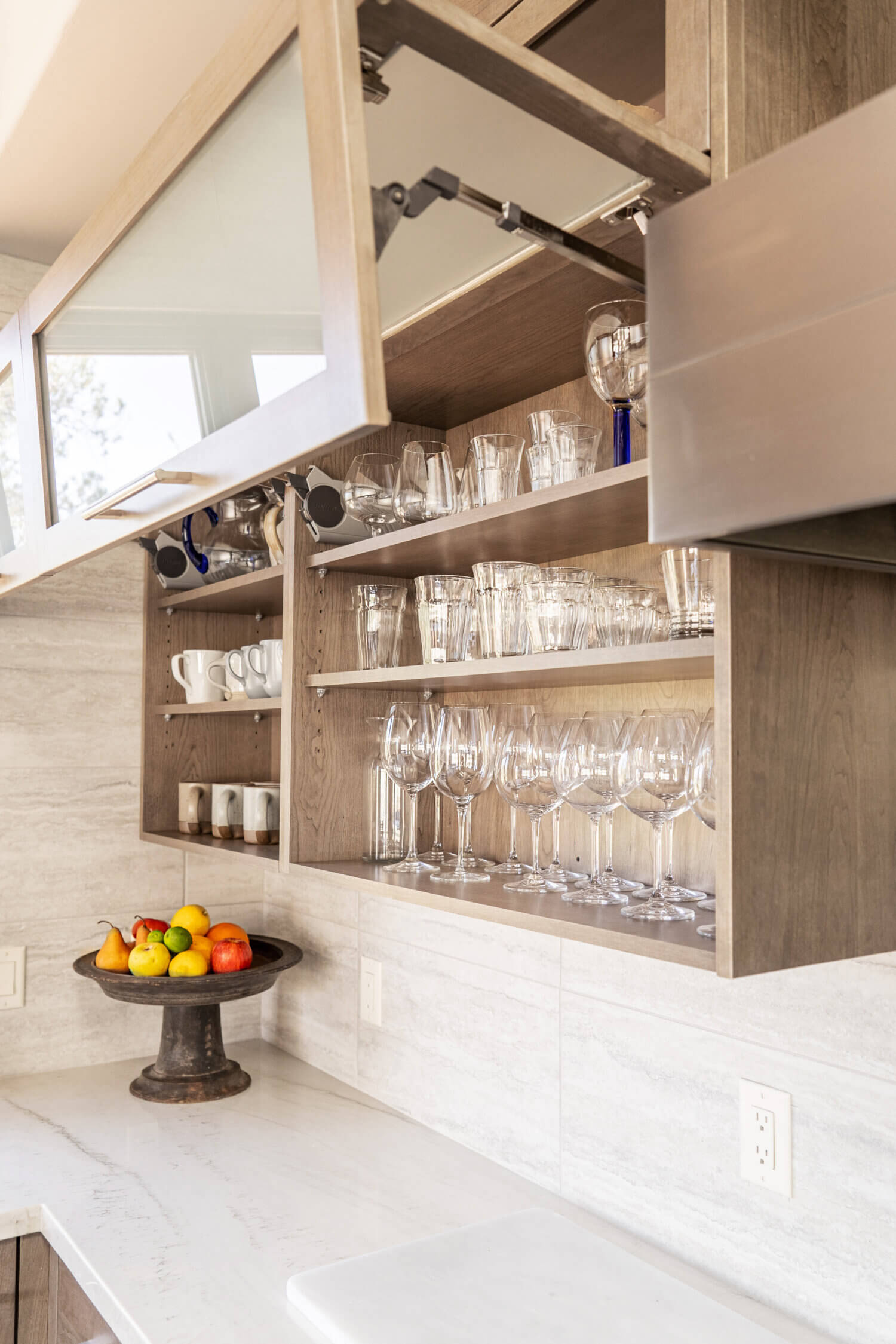 Frameless wall cabinets with lifting bi-fold glass cabinet doors for storing a large glassware collection in light gray stained cabinets from Dura Supreme.