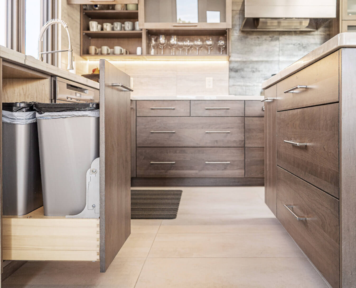 A close up of the base cabinets with a pulled-out base recycling cabinet with two waste bins and open wall storage with lifting cabinet door.