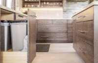 A close up of the base cabinets with a pulled-out base recycling cabinet with two waste bins and open wall storage with lifting cabinet door.