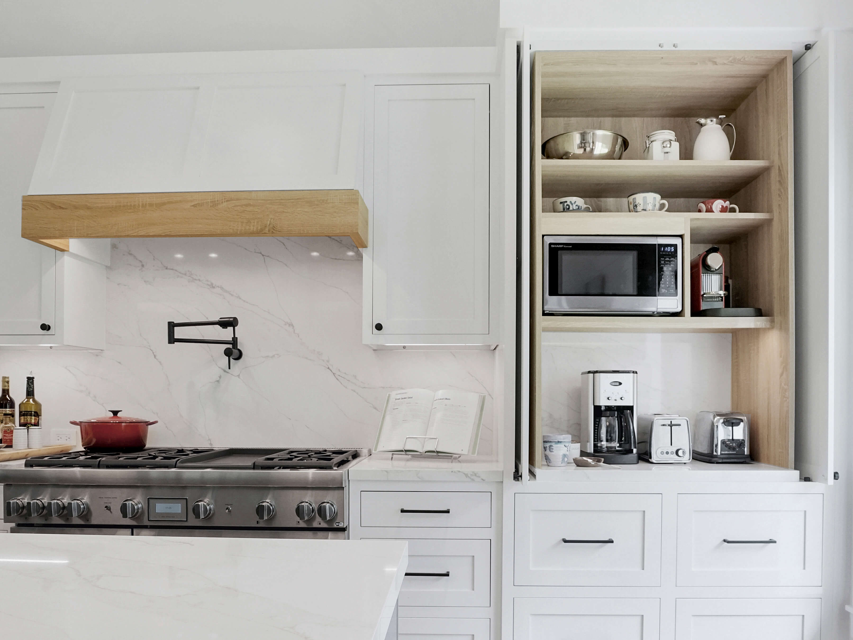 This beverage station hides all of the small appliances for crafting beverages as well as a toaster with an appliance door above the beverage fridge as well as cup storage in the cabinet to the side and pantry storage for coffee supplies and breakfast fixings in the cabinet above. The kitchen has white painted inset cabinets and a modern wood hood with a light stained wood frieze accent.