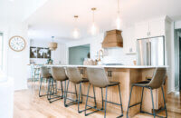 An angled view of the kitchen in an open floor plan with a light stained wood kitchen island and wood hood contrasted by white painted cabinets with shaker doors.