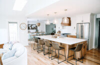 An angled view of the kitchen in an open floor plan with a light stained wood kitchen island and wood hood contrasted by white painted cabinets with shaker doors.