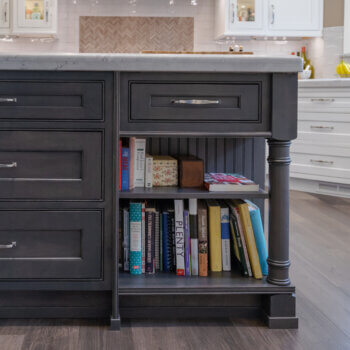 A table-like kitchen island end cap with a decorative turned post leg and shelves beneath a drawer for open storage. Shown in a dark gray stained wood with traditional styling.