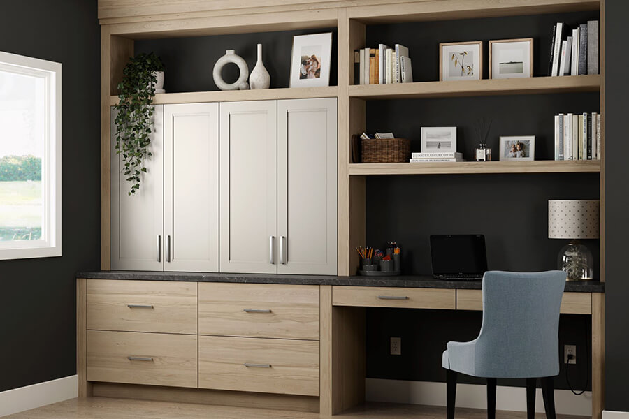 The Dura Supreme office cabinets and built-in desk feature a combination of a slab door style in light stain on the Hickory wood cabinets paired with a detailed flat panel door style in a natural off-white painted finish.