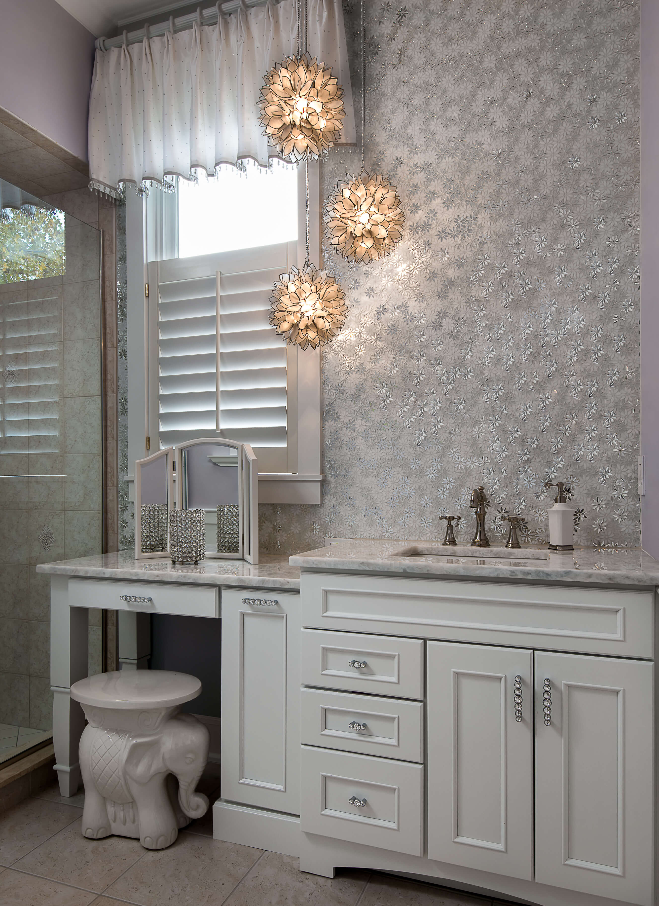 A glamourous powder room vanity with a makeup desk with white painted bathroom cabinets that have full overlay flat panel doors. A metallic wallpaper and dazzling pendent lights had a spark of glam to the remodeled room.