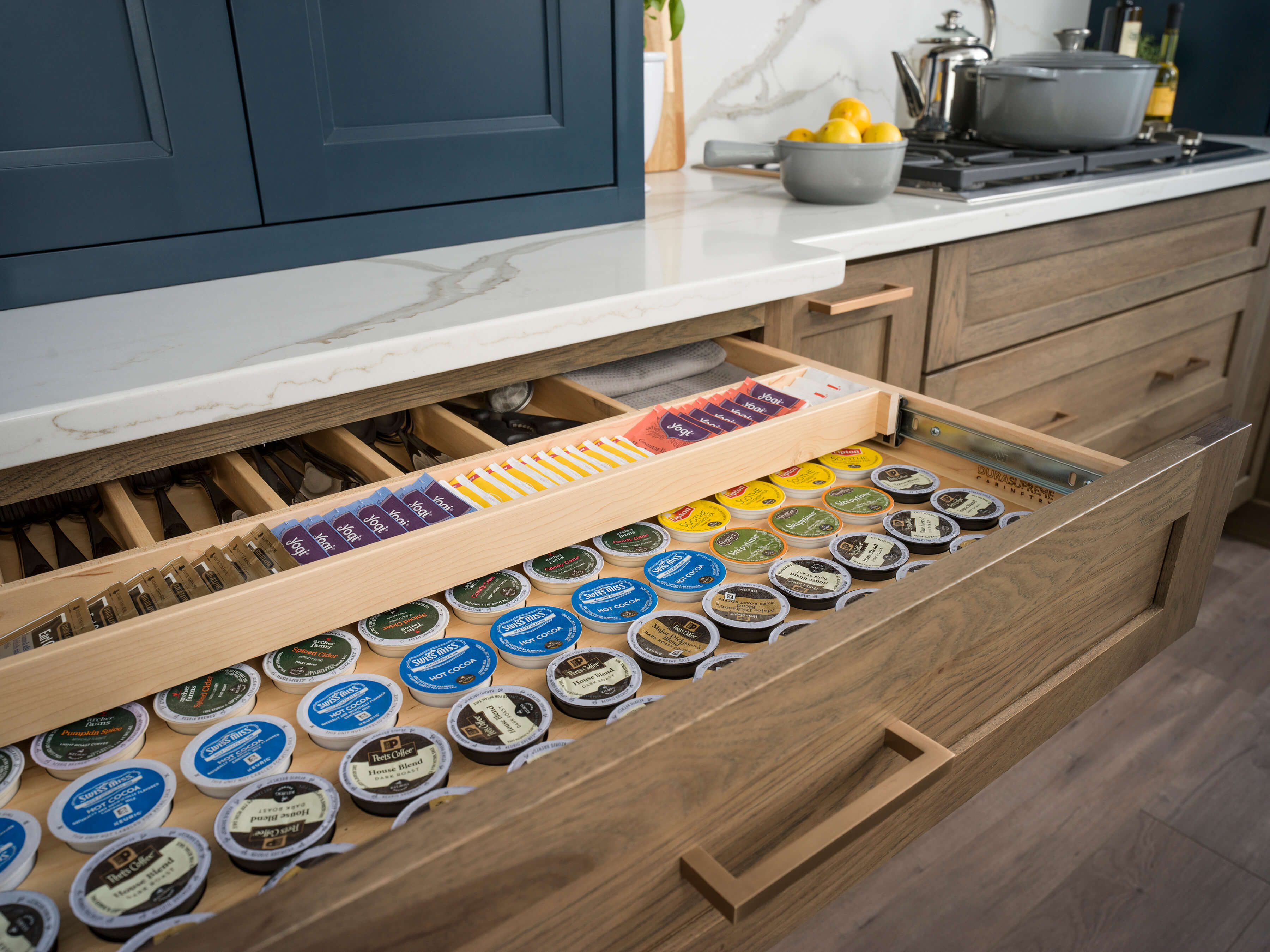 The base cabinet has a multi-functional two-tier drawer that provides storage for a silverware set and large collections of k-cups and tea bags. The lower cabinets are shown in a medium stained hickory wood with brushed brass cabinet hardware.