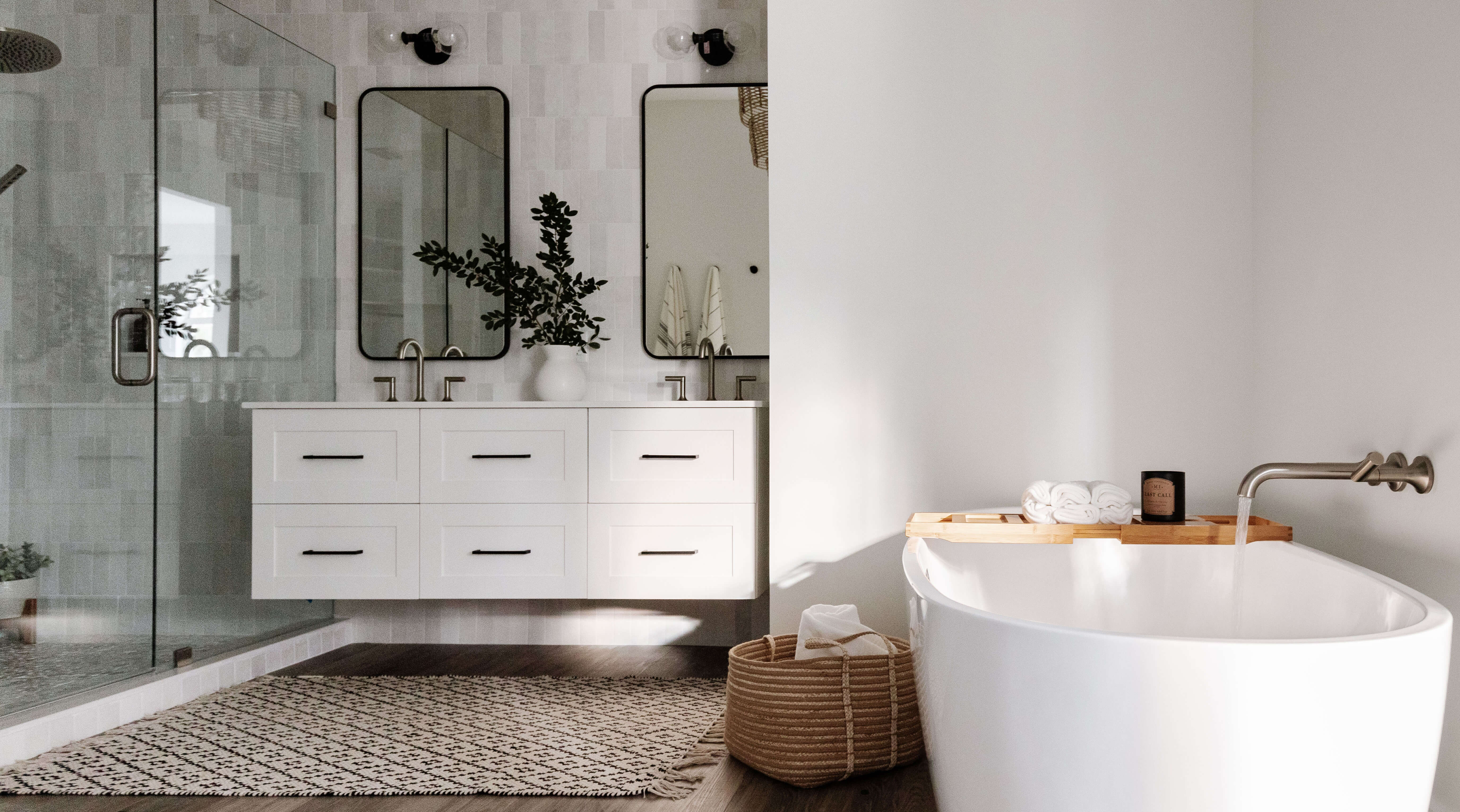 This master bathroom design features a white painted floating vanity with a pretty flat panel door style. The room has a soft, neutral color palette featuring light gray painted walls, lightly stained wood flooring, white and gray tiles, a white freestanding bathtub.