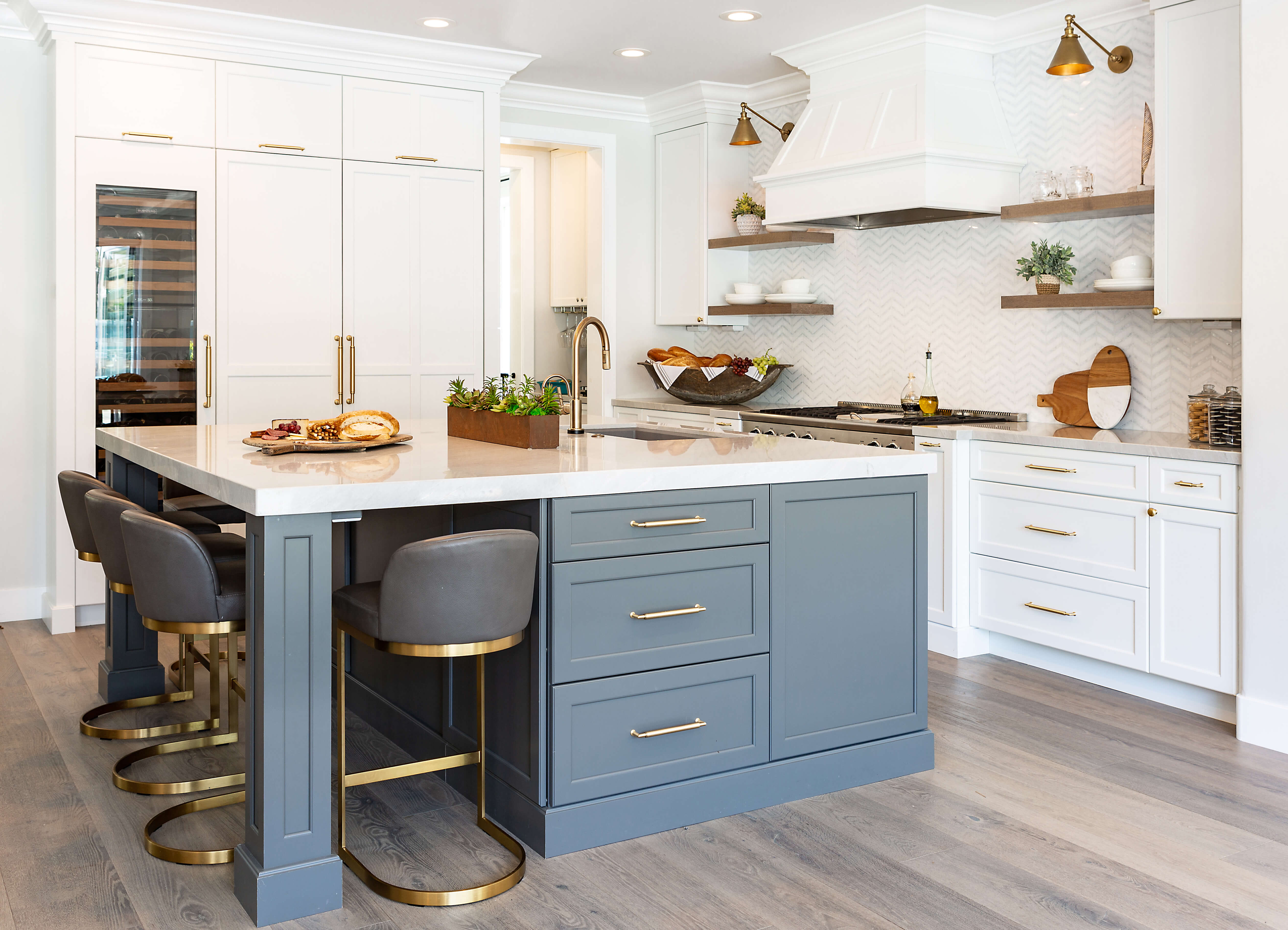 A two-tone transitional kitchen remodel with white painted frameless cabinetry and a medium gray painted kitchen island with seating and a kitchen island sink. The modern white painted wood hood is surrounded by wooden floating shelves. Frameless cabinets can be used for both contemporary and traditional door styles. Since the reveals are so tight between the doors/drawers, you will need to be sure there are adequate clearances for them to open and close properly.