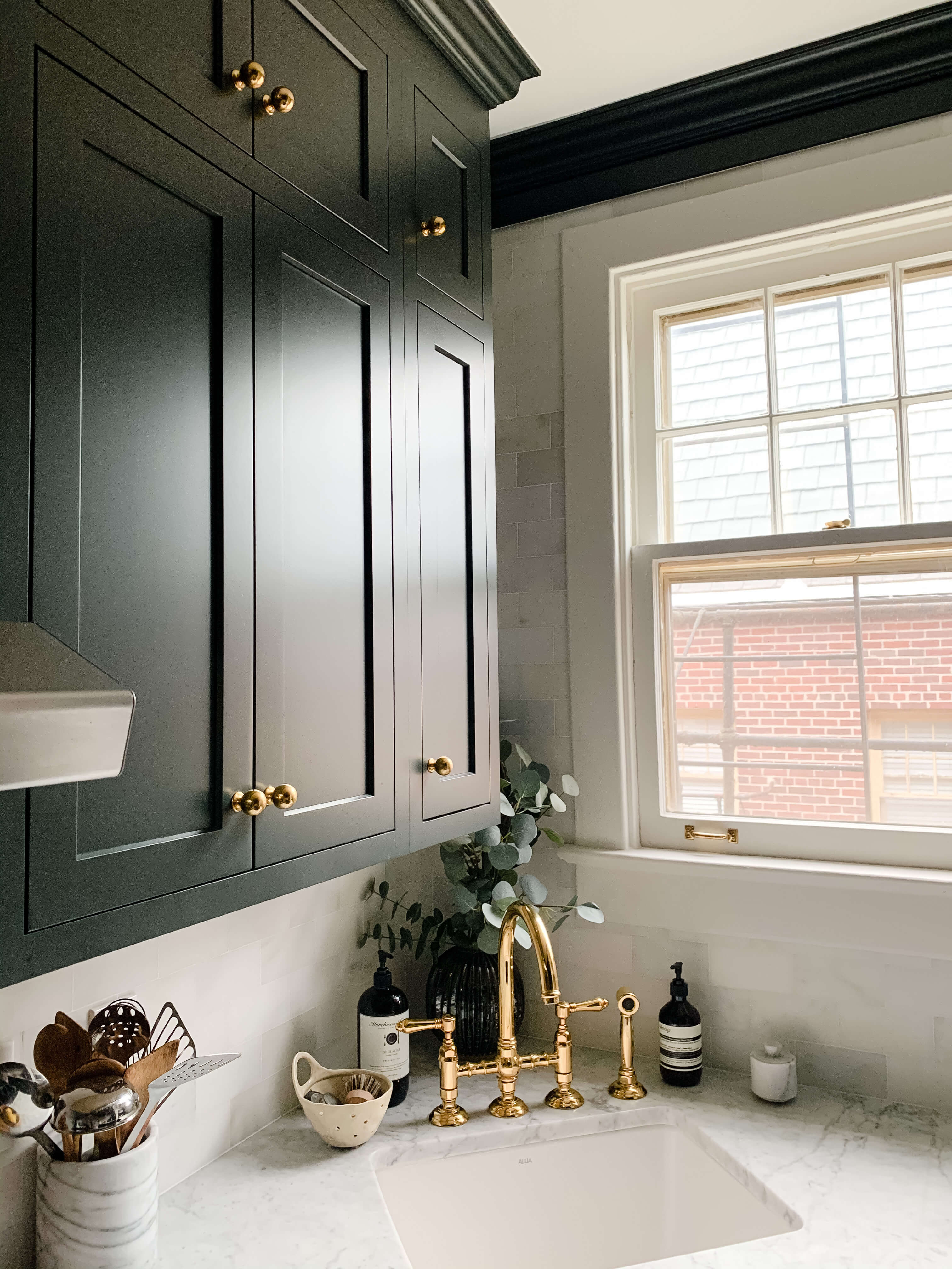A dark deep green painted kitchen with inset cabinet doors in an old historic styled home. A cabinet knobs aand the corner kitchen sink faucet features brassy gold finishes to accent the dark green color palette. .