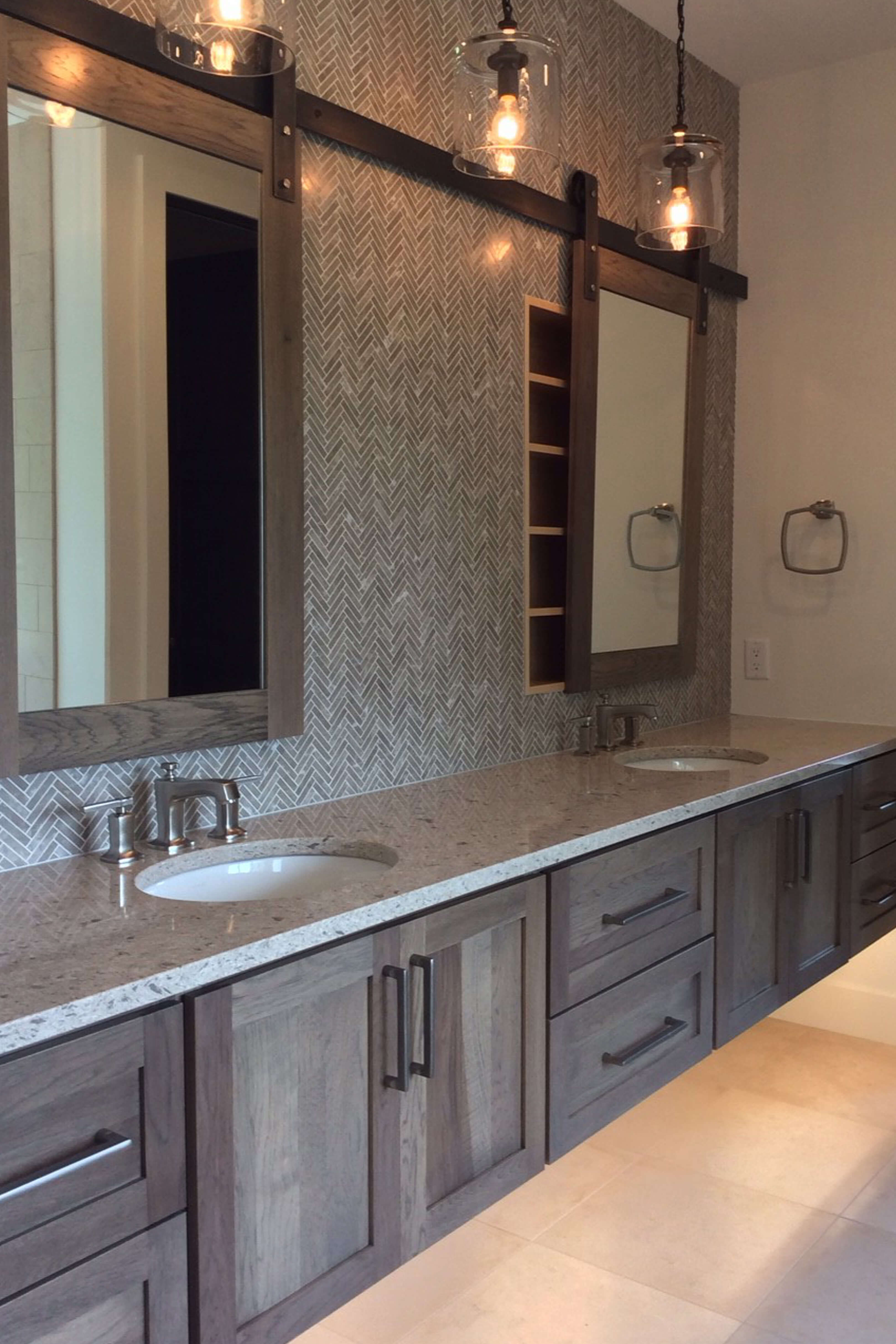 A modern rustic floating vanity cabinet with double sinks using full overlay shaker door styles in a gray stained Hickory wood. Barn door style hardware is used for the medicine cabinet mirrors to create sliding mirror doors.