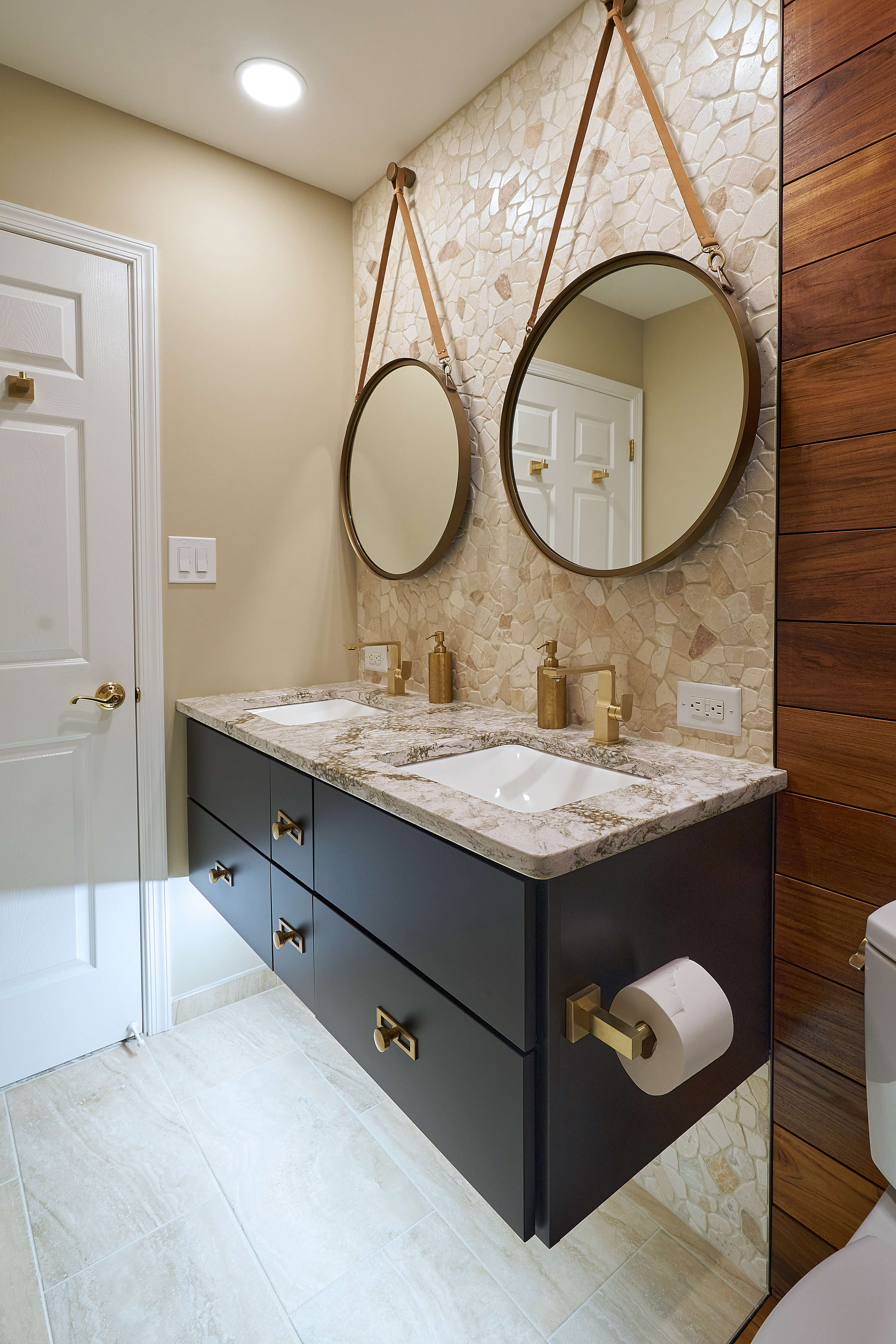 A contemporary bathroom design with warm stone and wood wall paneling featuring a modern black painted wall hung vanity with two bathroom sinks and two round rope-hanging vanity mirrors.