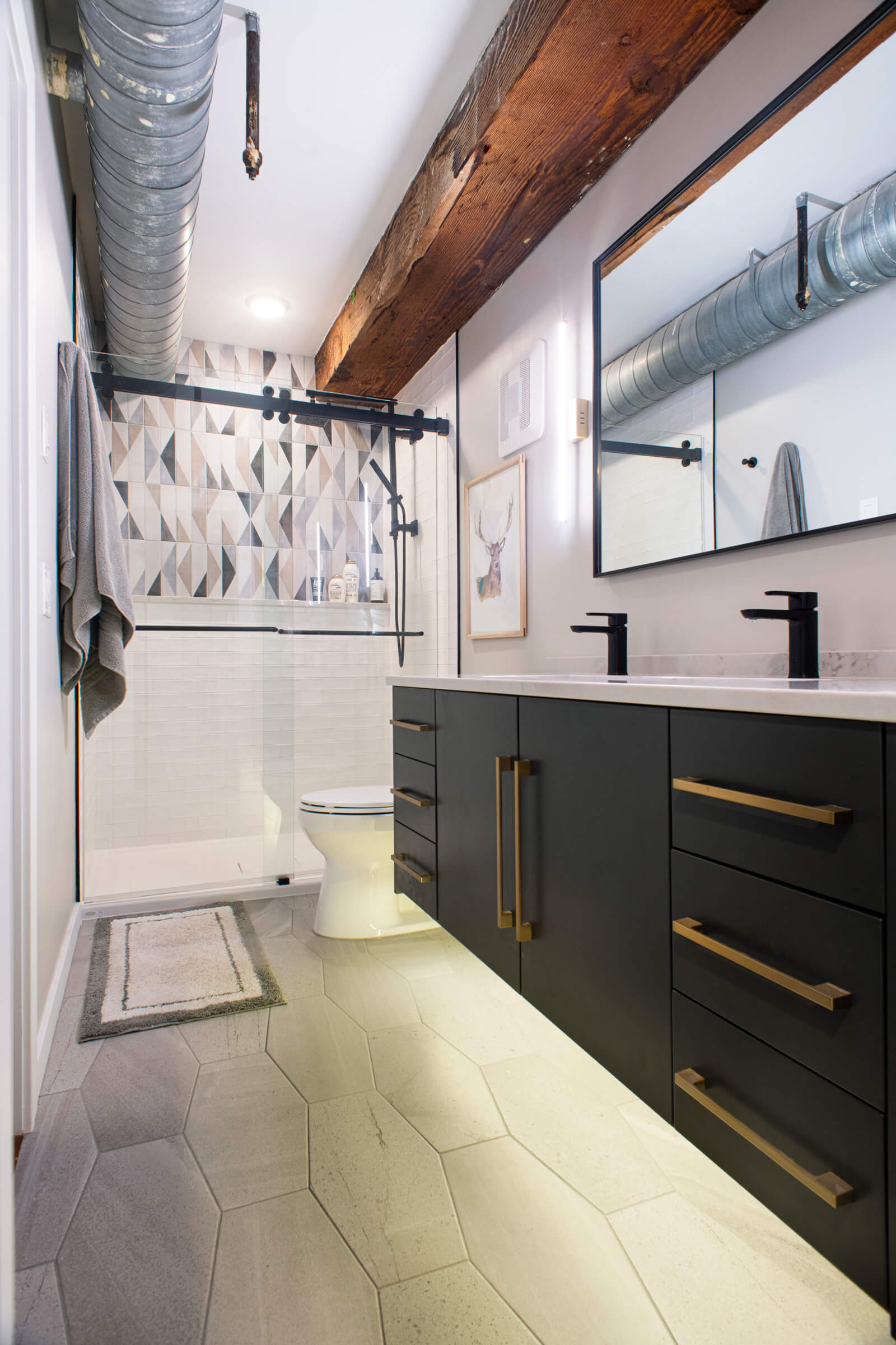 An industrial styled master bathroom design with dual sinks in a black finish, a white vanity countertop, and black painted floating bath cabinets with brushed brass hardware pulls. An industrial-like air duck pipe and a railroad tie-inspired ceiling beam add to the industrial style of the bathroom design.