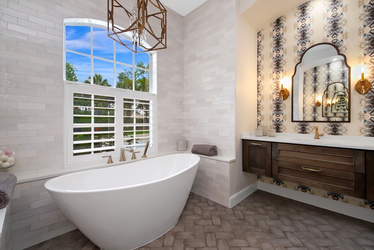 A glam style master bathroom with a dark stained wood vanity that is mounted to the wall for a floating effect. A delightful wallpaper pattern creates an accent wall behind the vanity accenting the floating vanity and vanity mirror area. A freestanding tub sits in front of a large window with a beautiful view with a dramatic chandelier hanging above.