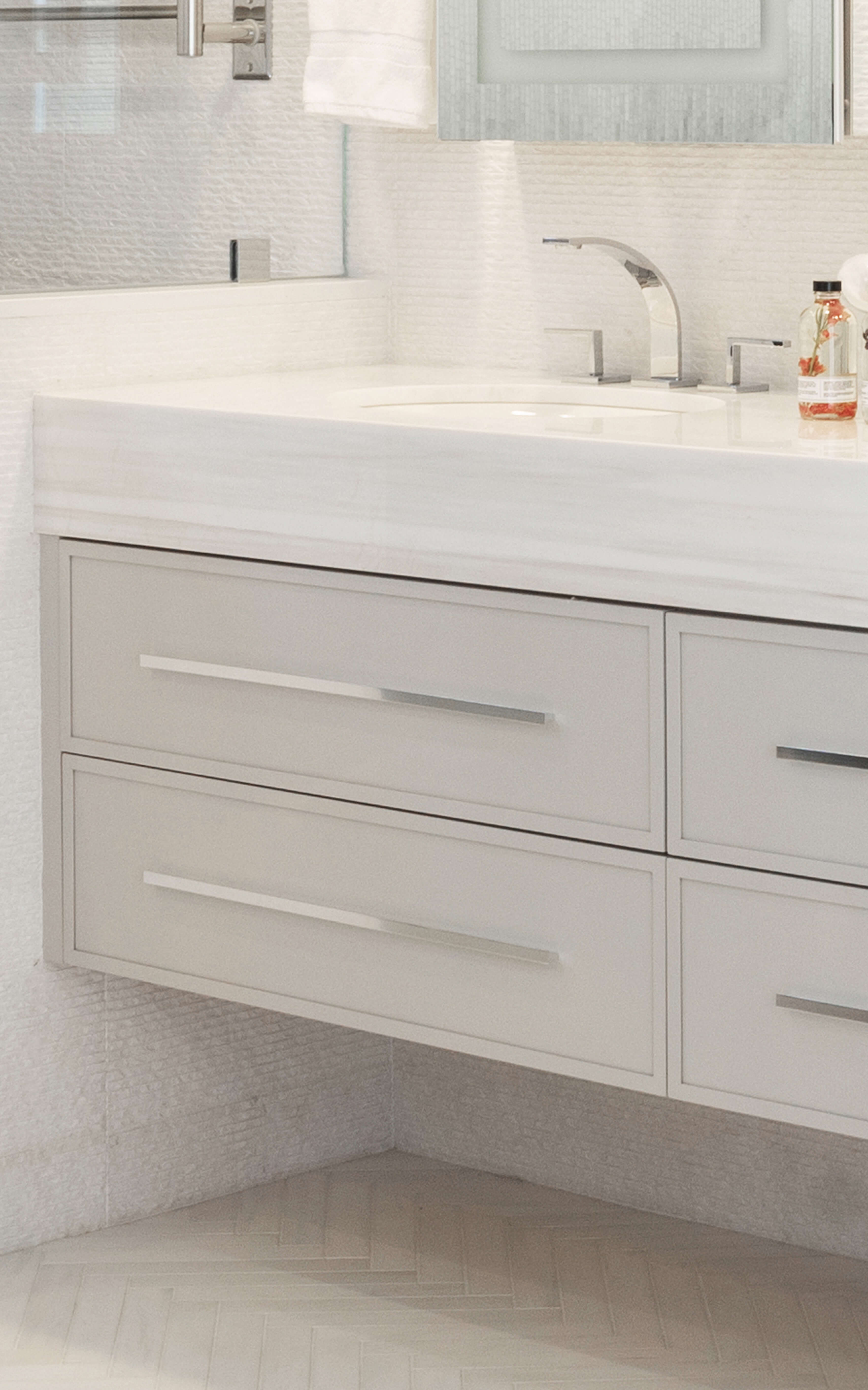 A close-up of the floating vanity with very thin profiles on the flat panel door. The wall hug vanity is shown with a soft, light gray painted finish, long satin nickel hardware pulls, and a thick quartz countertop.