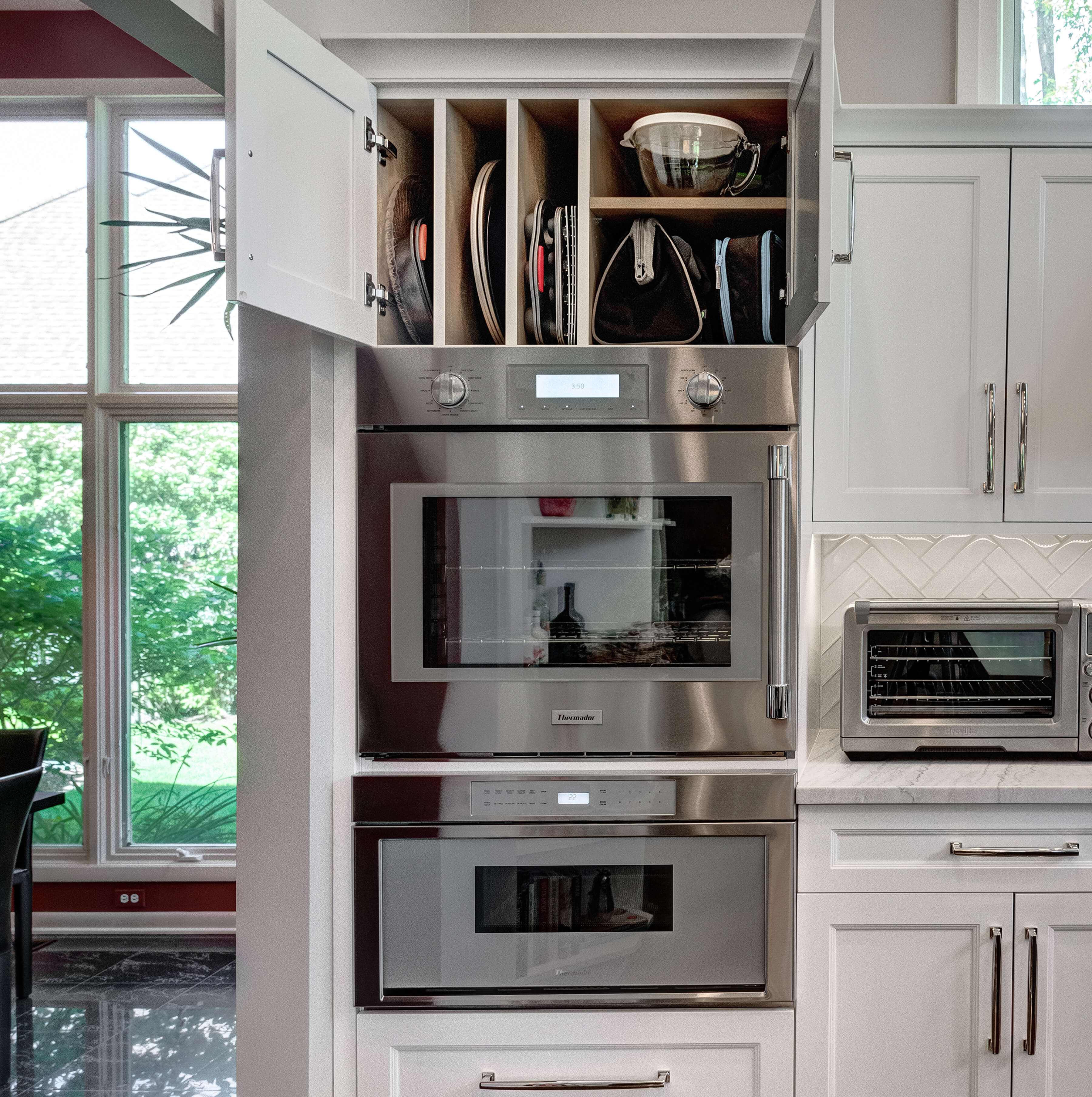 An all-white kitchen design with kitchen storage above the wall ovens using partitions for tray and pan storage and a shelf for organized bakeware containers.