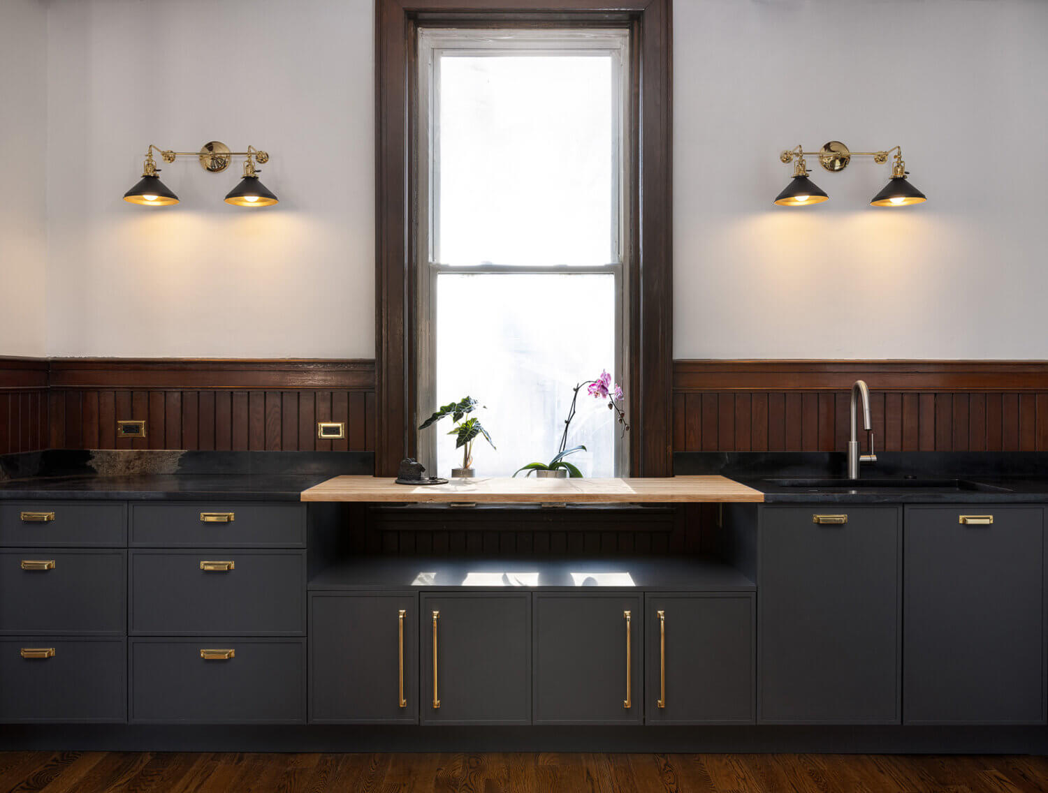 A wall with only base cabinets in a dark charcoal gray paint with skinny shaker door styles with a shelf across the window.