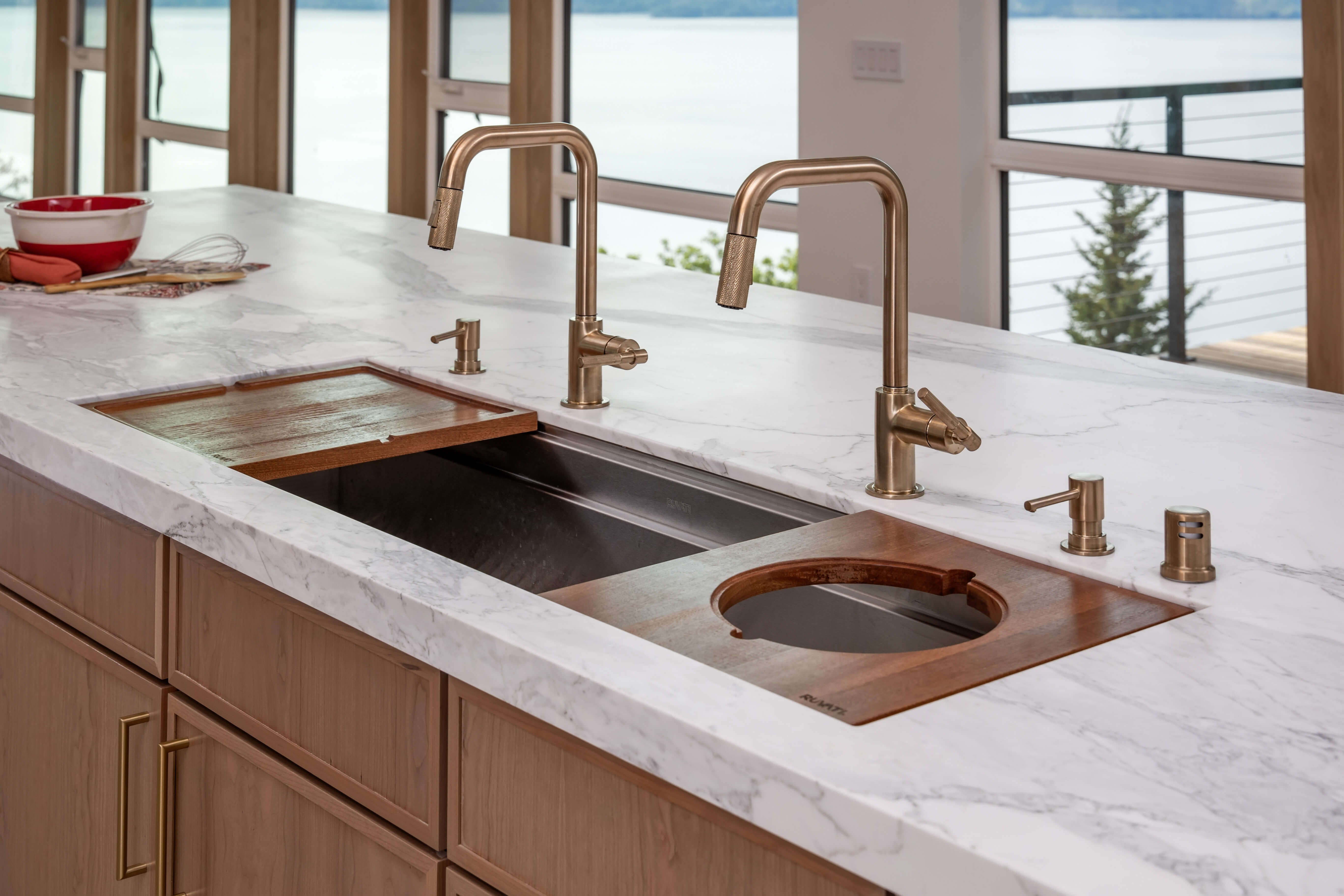 A large kitchen sink and clean-up workspace with dual kitchen faucets in a brushed brass finish with warm stained wooden cutting boards.