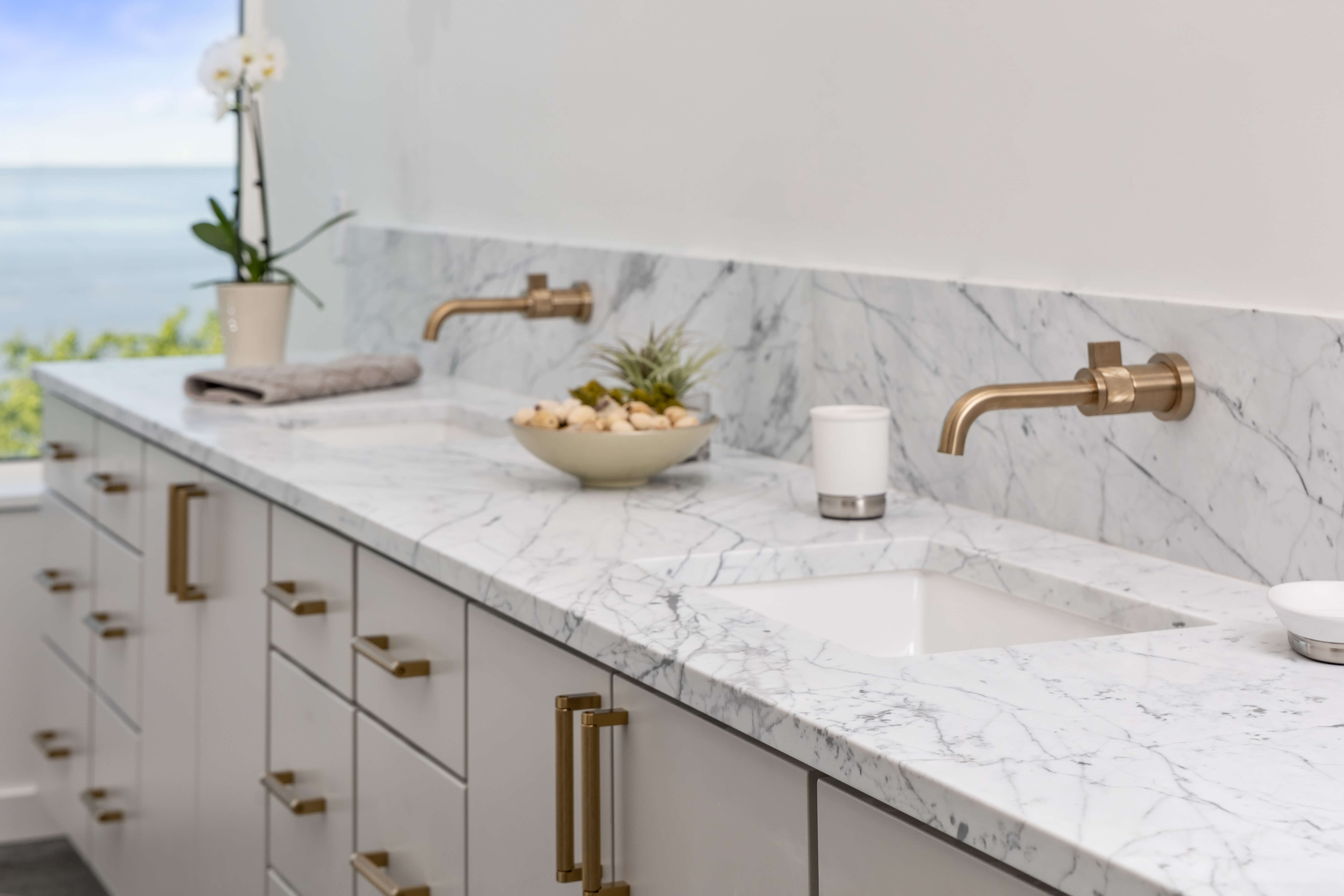 A soft, light gray bathroom vanity with slab cabinet doors, dual sinks, and brushed brass hardware accents.