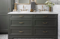 A dark green (almost black) painted bathroom vanity and tall linen cabinet with brass hardware and light gray tiles.