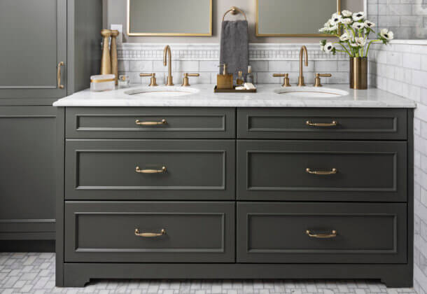 A dark green (almost black) painted bathroom vanity and tall linen cabinet with brass hardware and light gray tiles.