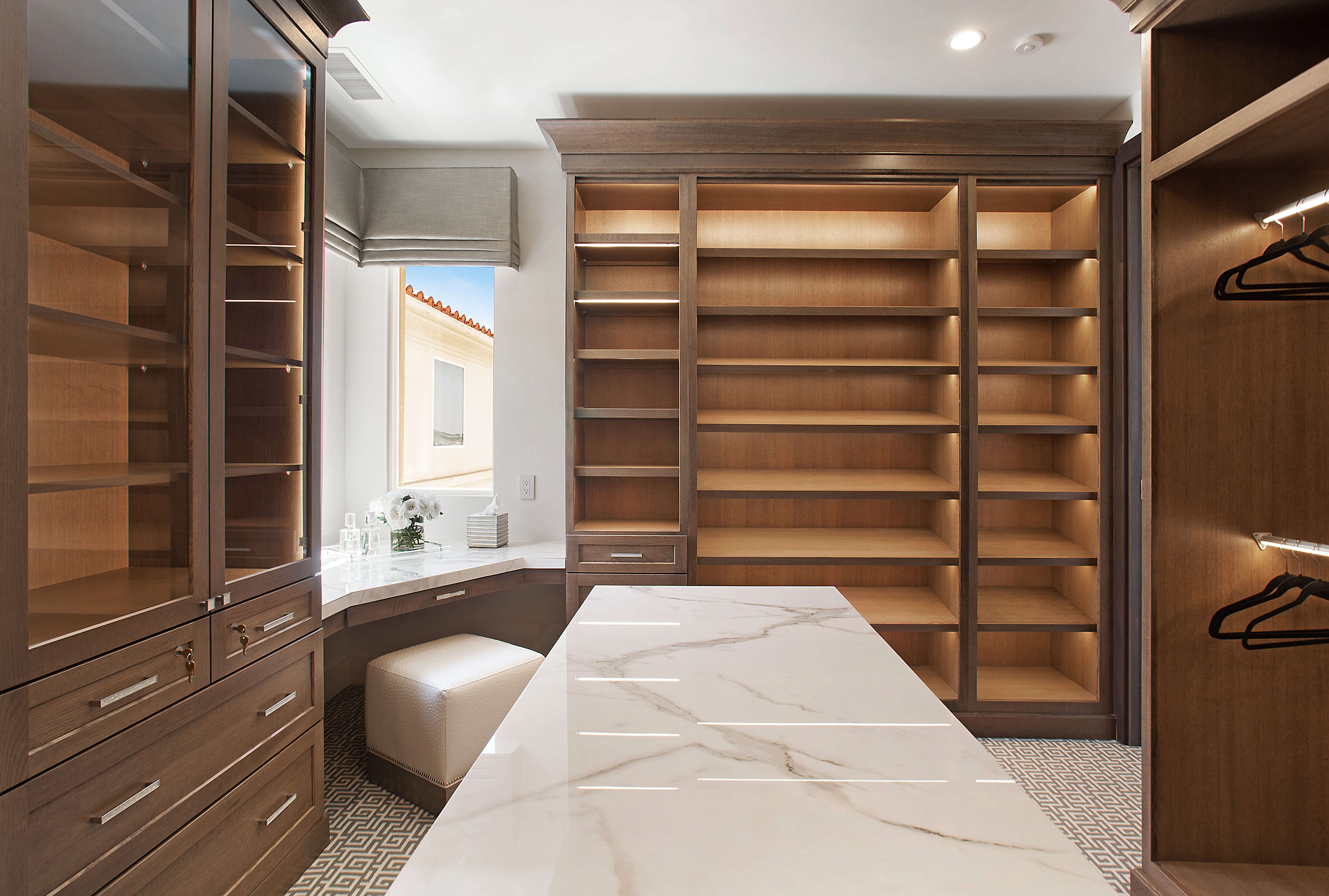 A beautiful master bedroom closet design with 3 walls of custom storage, a island in the center, and a corner beauty desk with seating.