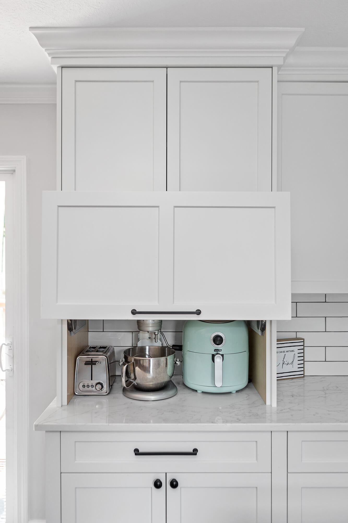 A larder cabinet with a lift-up cabinet door that hides several powered appliances at countertop level. Shown open.