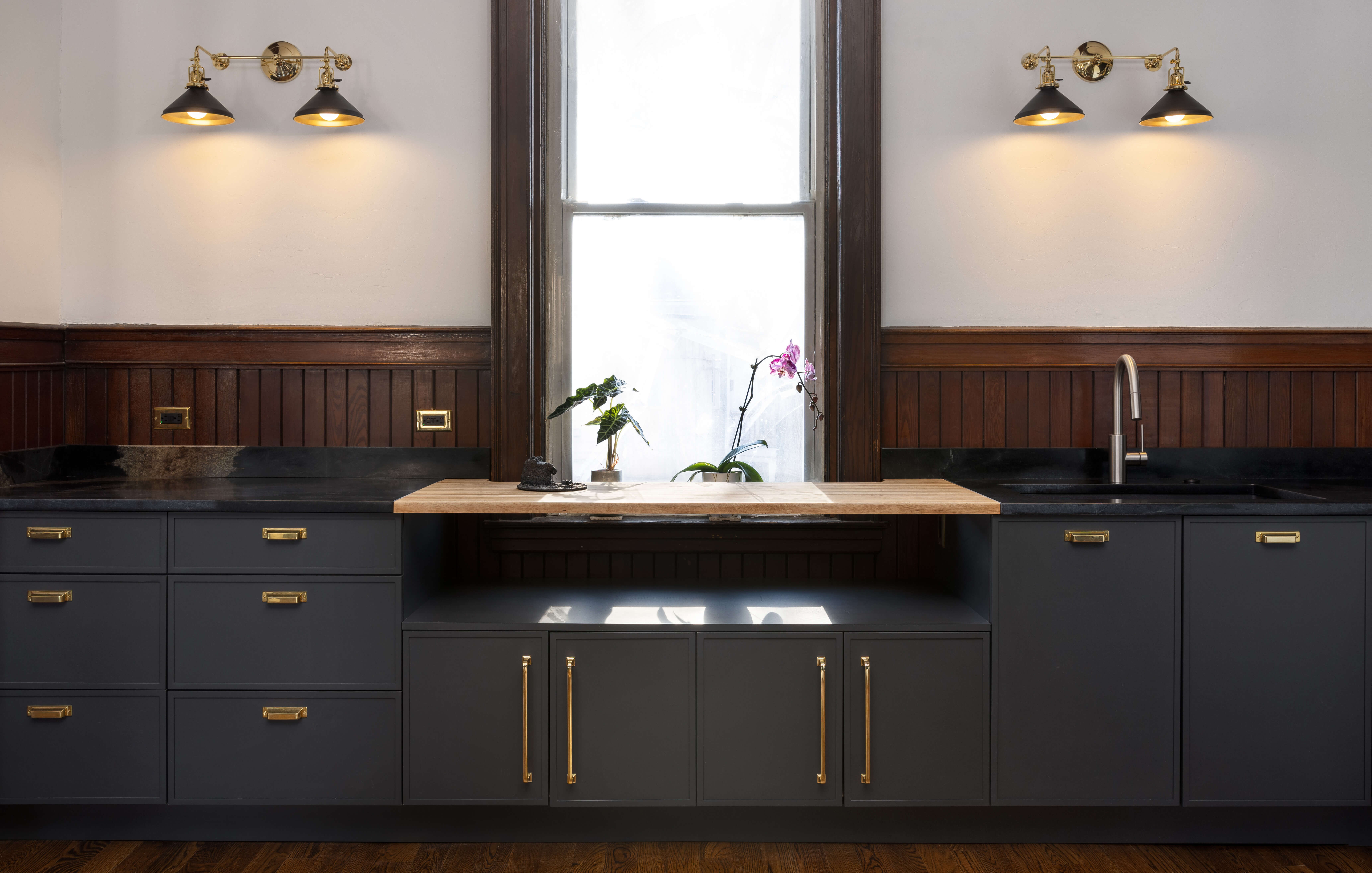 A wall with only base cabinets in a dark charcoal gray paint with skinny shaker door styles with a shelf across the window.