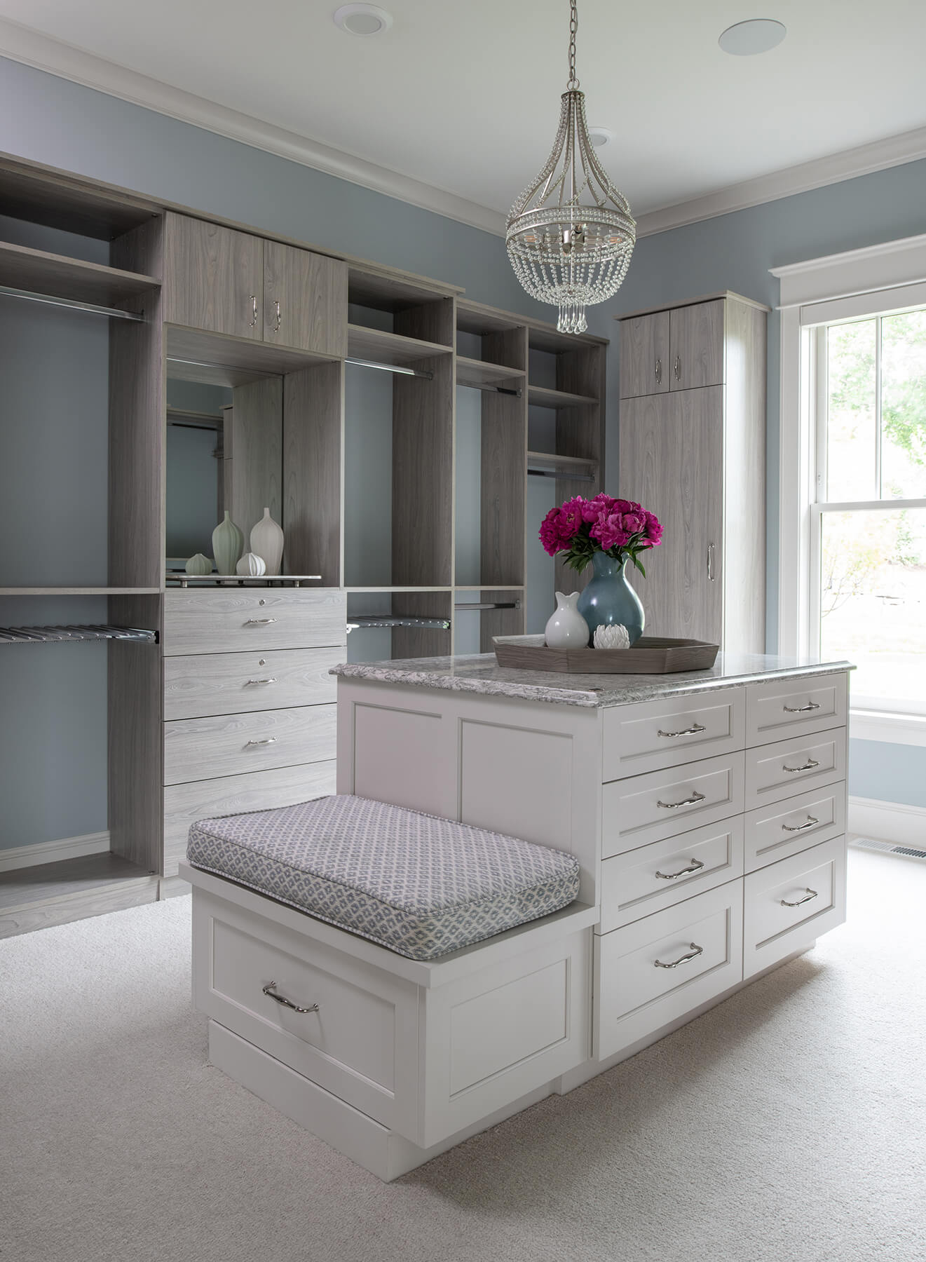 A bright and inviting walk-in closet design with custom cabinetry from Dura Supreme in a light gray texture with a white painted closet island with a built-in boot bench.