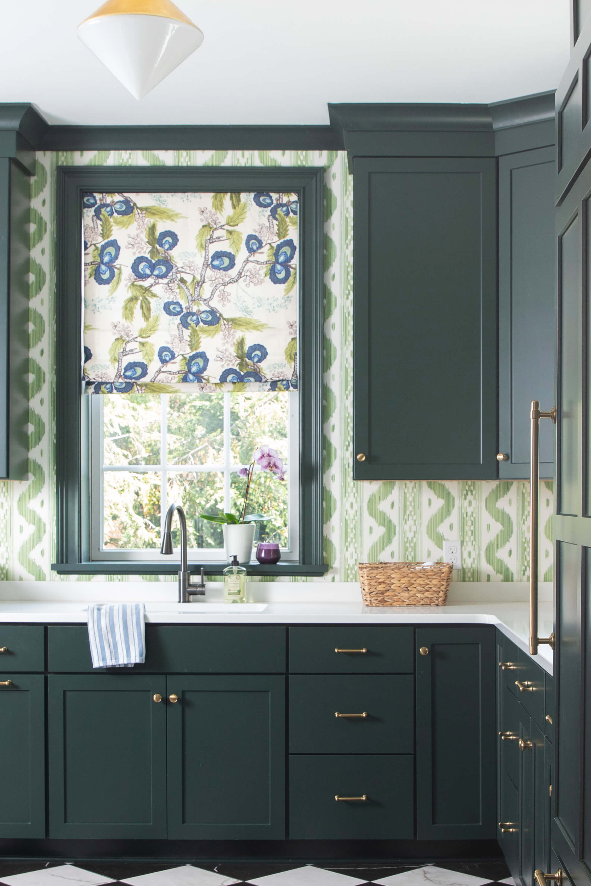 A colorful kitchen design with vibrant green and white wallpaper, dark green painted cabinets & trim and bright white countertops.