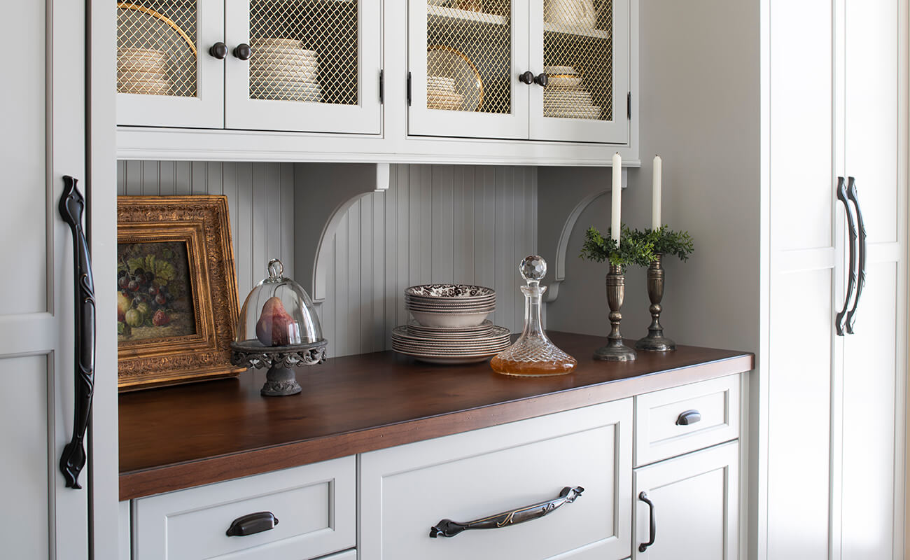 An English styled kitchen with light gray painted cabinets, wire mesh cabinet inserts, and a rich wood top.